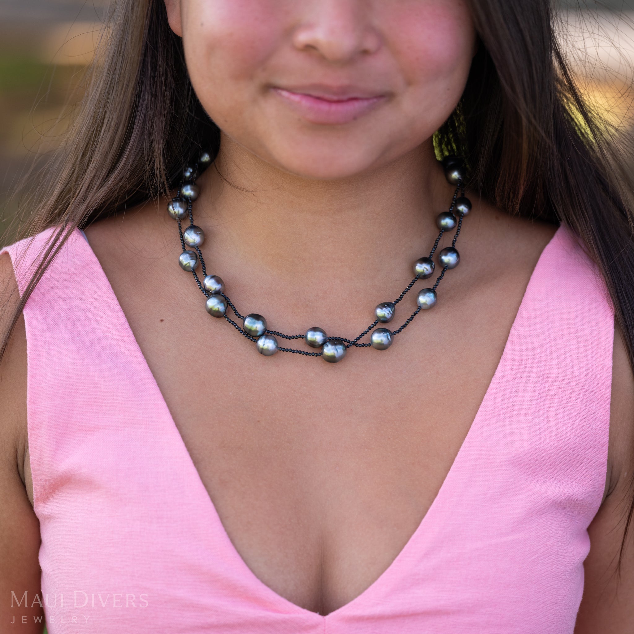 A close-up of a smiling woman in a light pink top wearing a Tahitian Black Pearl and Black Spinel Layered Necklace with a 14k yellow gold clasp, against a blurred background