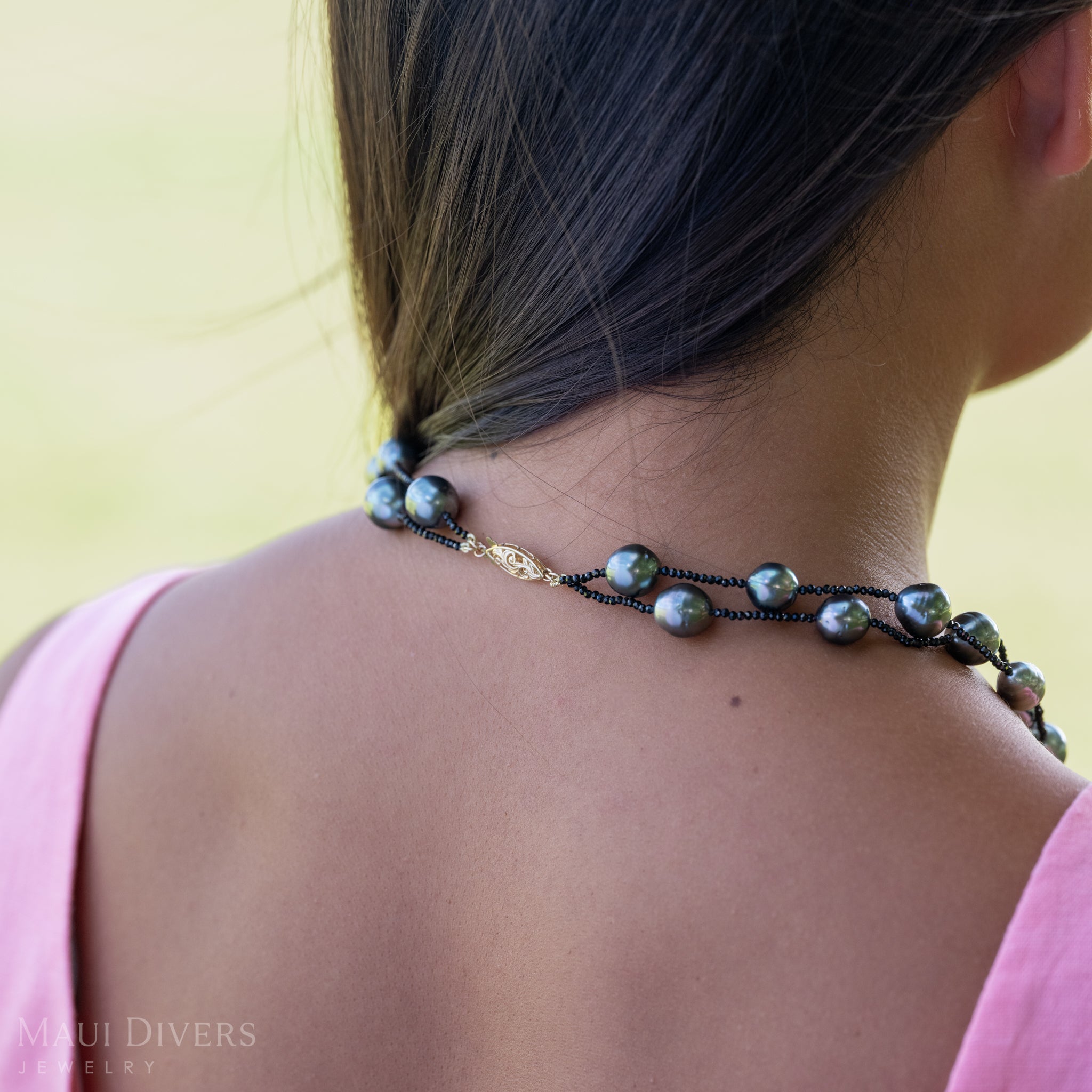 A close-up of the back of a woman in a light pink top showing the 14k yellow gold clasp of a Tahitian Black Pearl and Black Spinel Layered Necklace around her neck, against a blurred green grass background