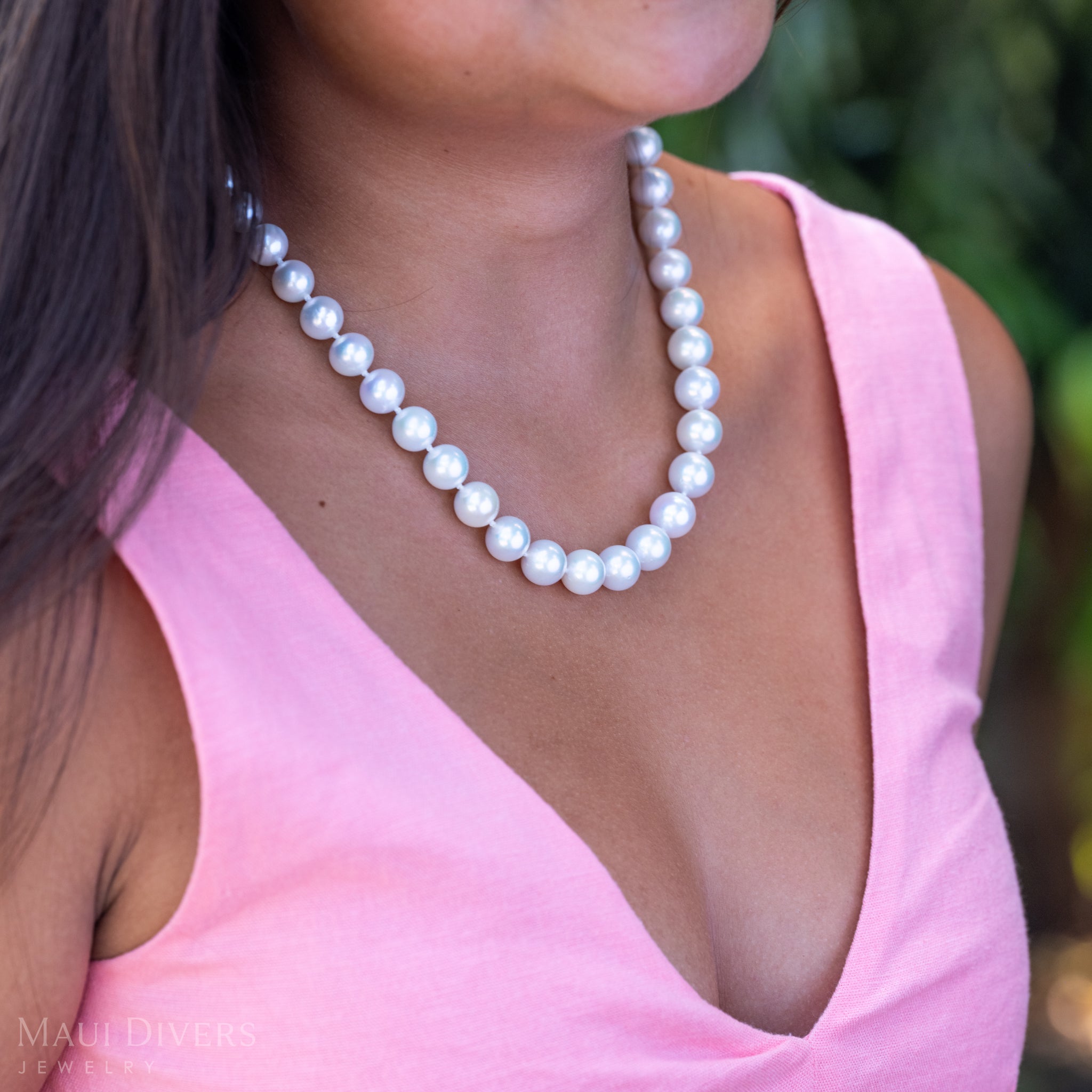A close-up of a woman in a light pink top wearing a Freshwater White Pearl Strand with Diamond Plumeria Clasp in 14k Yellow Gold, against a blurred leafy green background