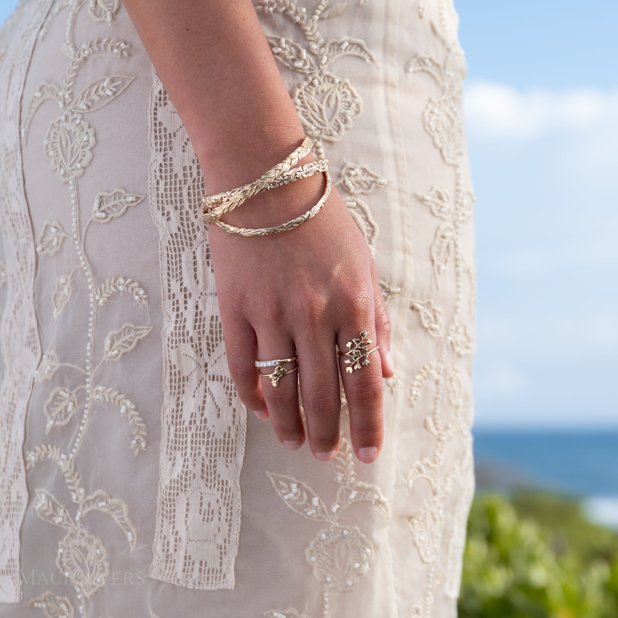 A close-up of a person's hand and wrist wearing 14k yellow gold Maiden Hair rings, a diamond anniversary ring, and stacked gold Hawaiian Heirloom bracelets, with a cream embroidered dress and a blurred outdoor background of greenery and blue sky.