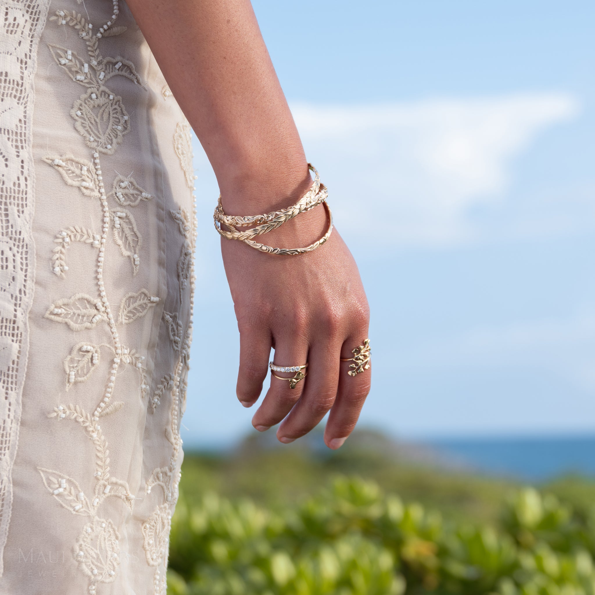 A close-up of a person's hand and wrist wearing 14k yellow gold Maiden Hair rings, a diamond anniversary ring, and stacked gold Hawaiian Heirloom bracelets, with a cream embroidered dress and a blurred outdoor background of greenery and blue sky.