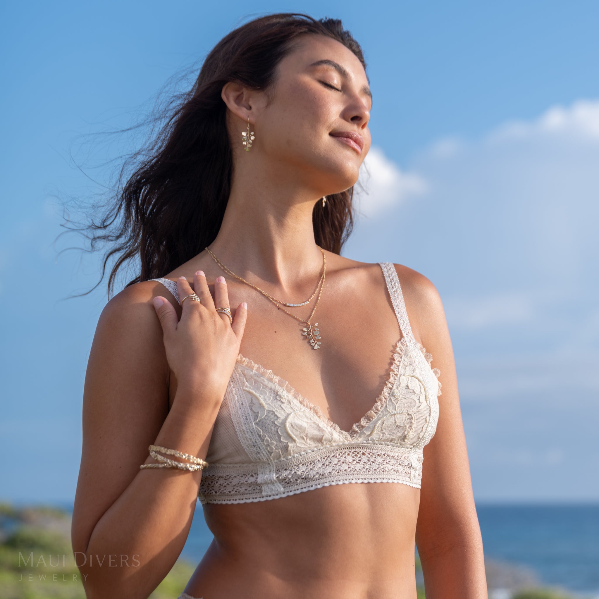 Woman wearing a gold Maiden hair fern pendant on a cable chain, maiden hair earrings, a Hidden Hawaiʻi lab-grown diamond necklace, and a lace top with a scenic background