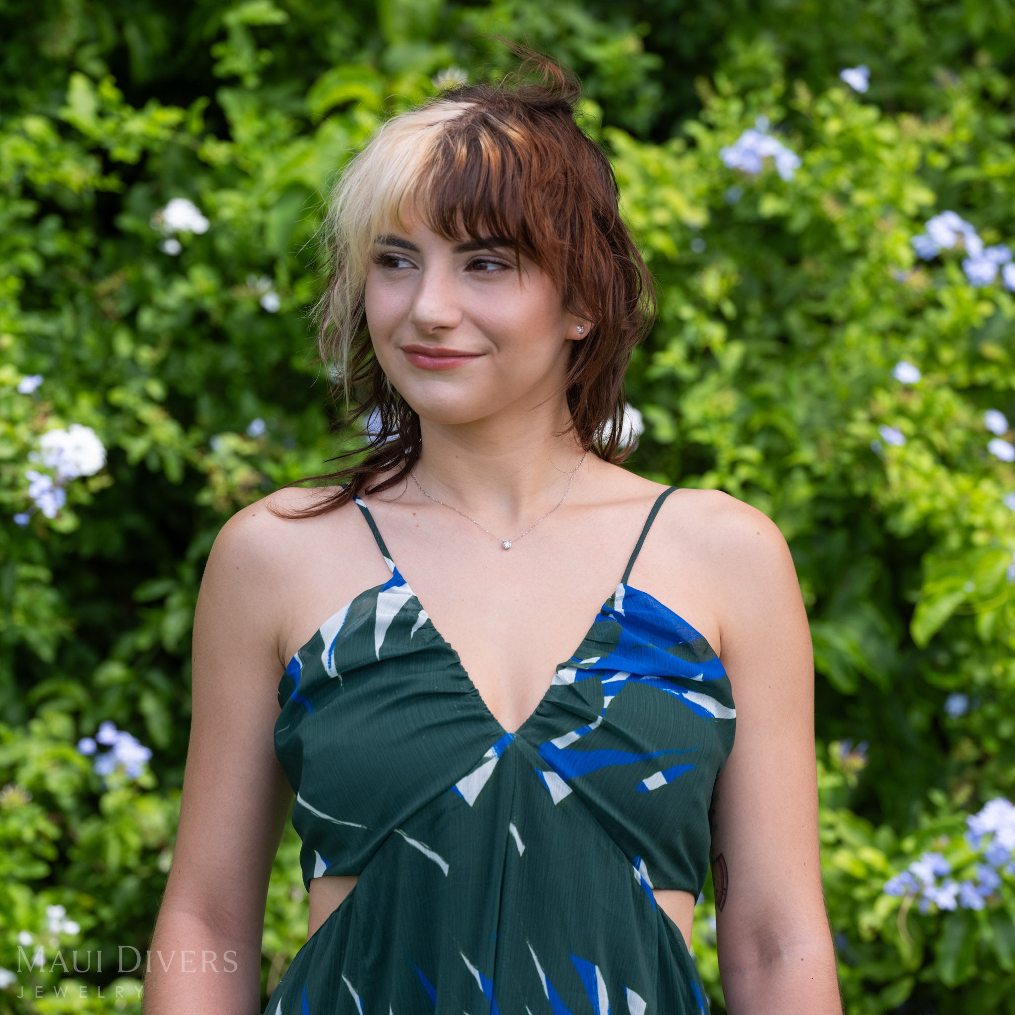 A lifestyle shot of a woman wearing a lab-grown diamond necklace with maile leaf prongs in 14k white gold and matching stud earrings against a blurry floral background