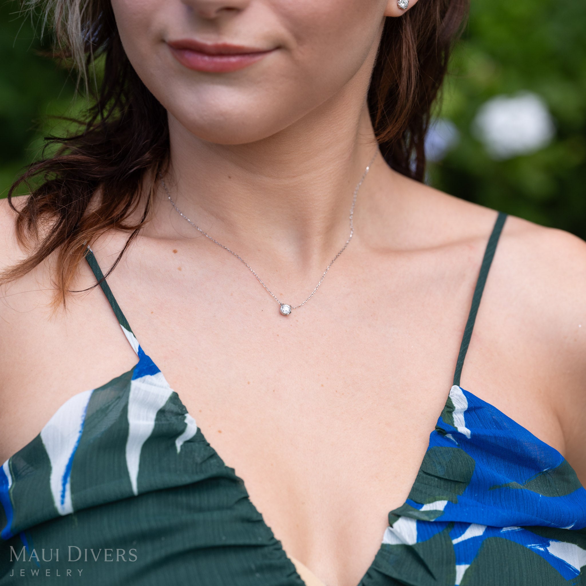 A close-up lifestyle shot of a woman wearing a lab-grown diamond necklace with maile leaf prongs in 14k white gold. The necklace is shown on her neck, with her blue and green patterned top and blurred green outdoor background visible.