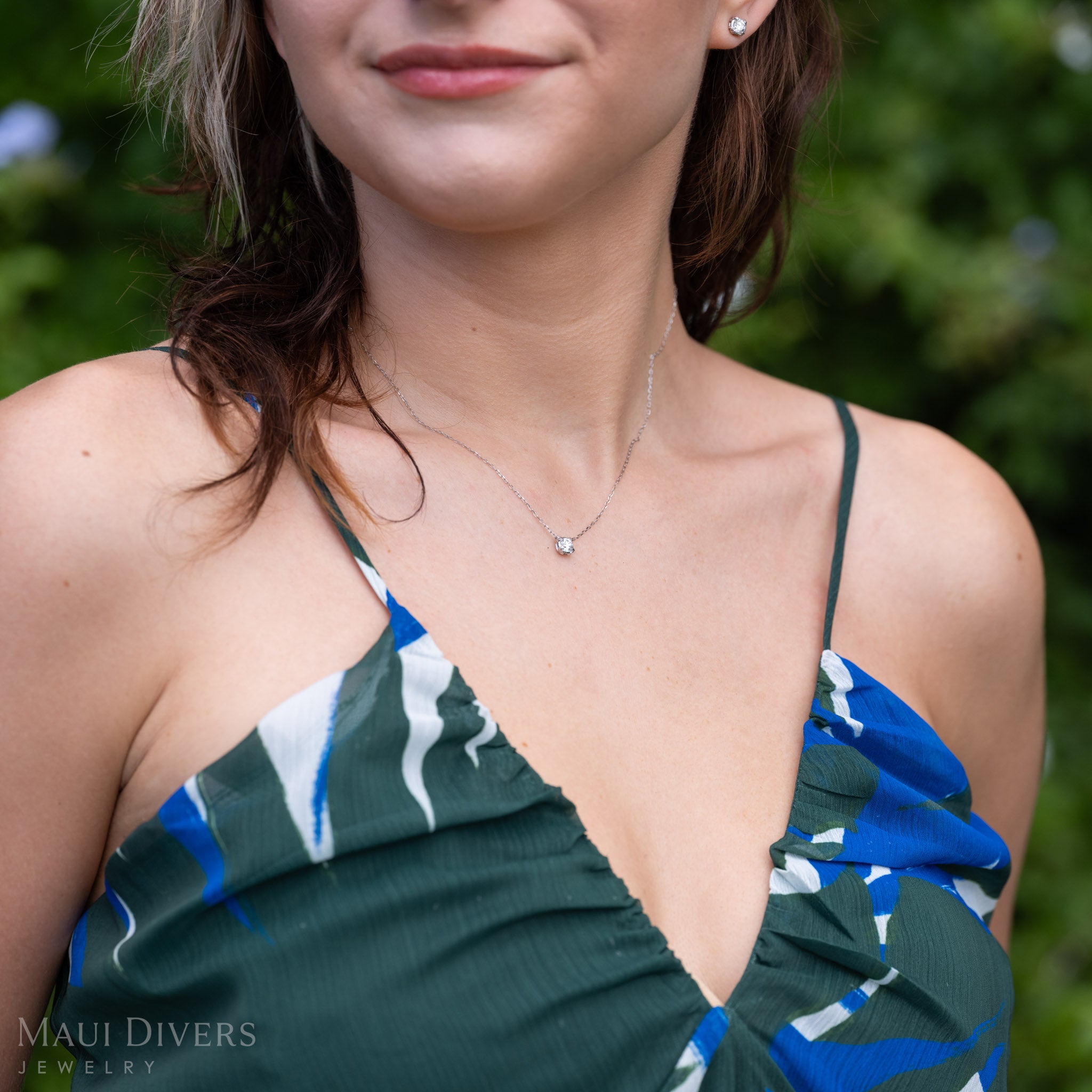 A close-up lifestyle shot of a woman wearing a lab-grown diamond necklace with maile leaf prongs in 14k white gold. The necklace is shown on her neck, with her blue and green patterned top and blurred green outdoor background visible.