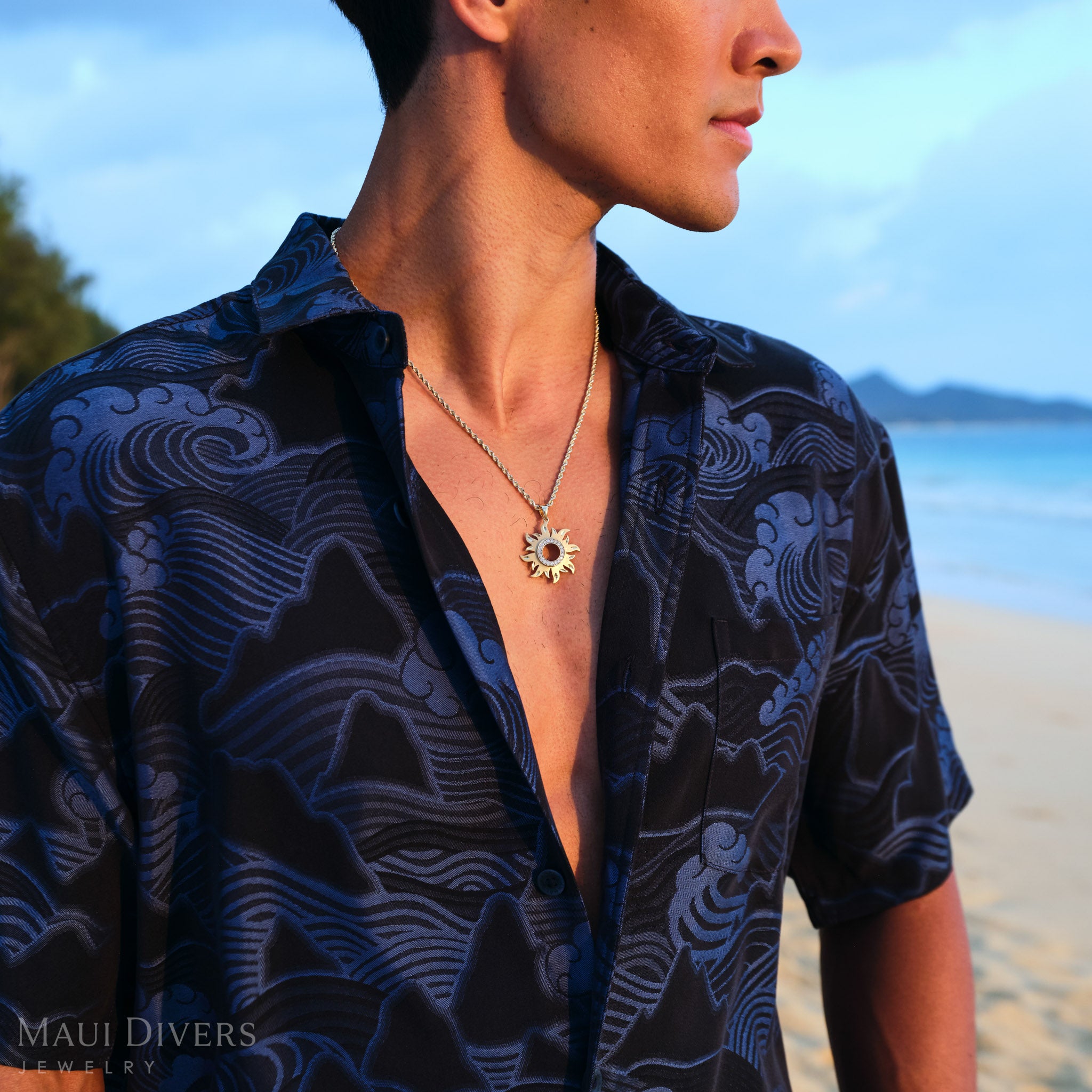 Man on a beach wearing a dark blue patterned shirt with a gold sun pendant on a rope chain