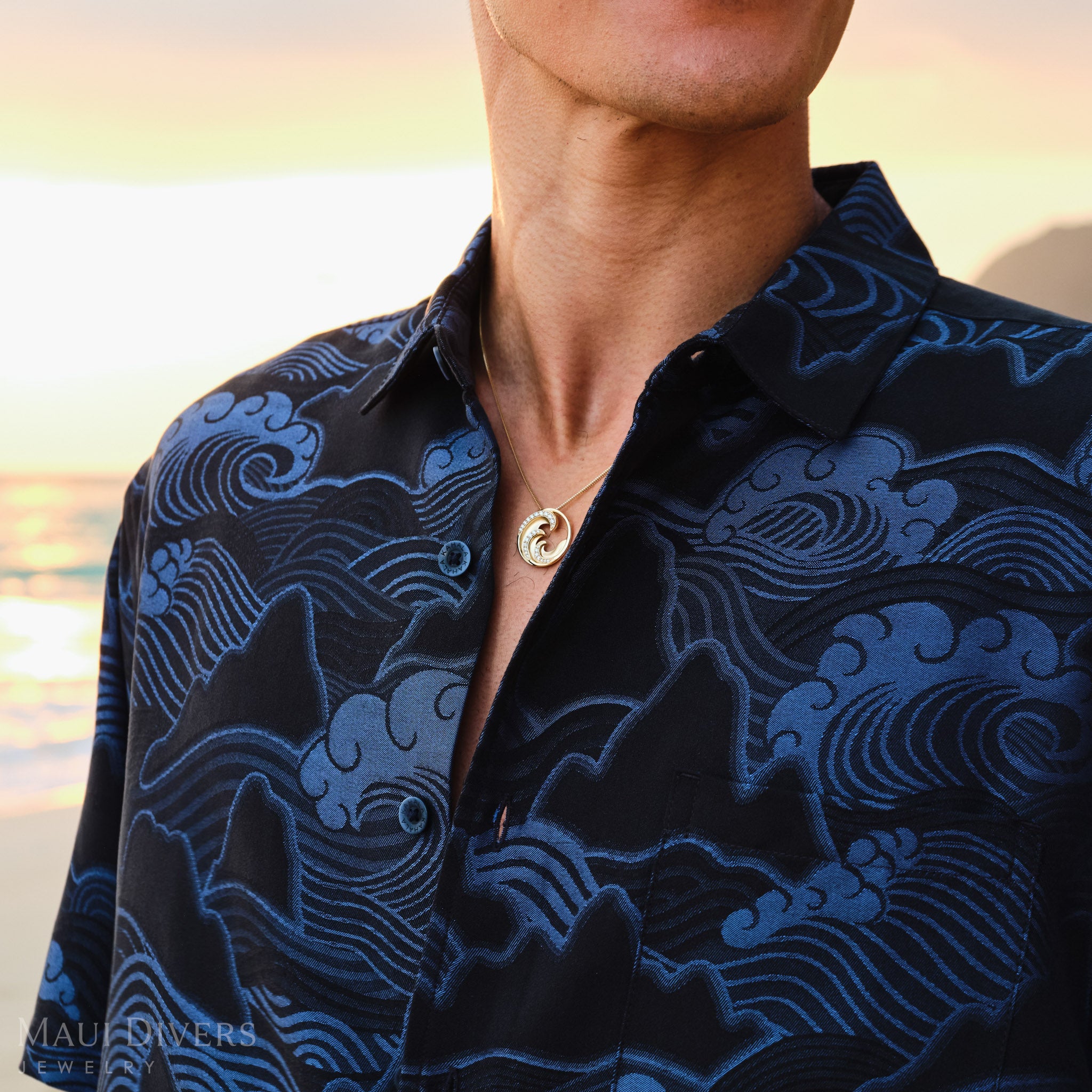 Man wearing a dark blue patterned shirt with a gold diamond Nalue wave pendant with a blurred sunrise background