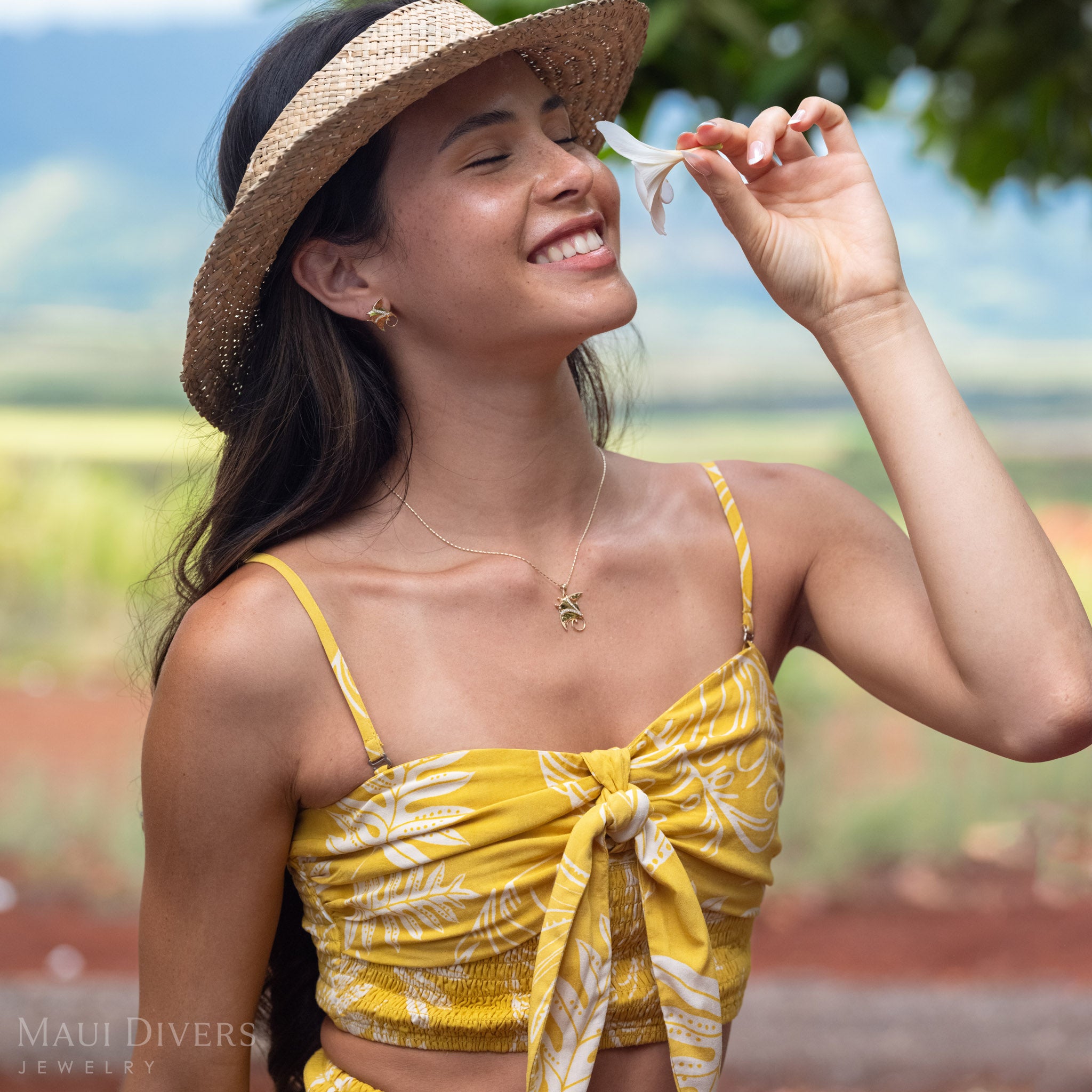 Woman smelling a white plumeria flower wearing a 14k Gold Manta Ray Earrings with Natural Diamond detailing and a matching necklace, wearing a brown woven hat. 