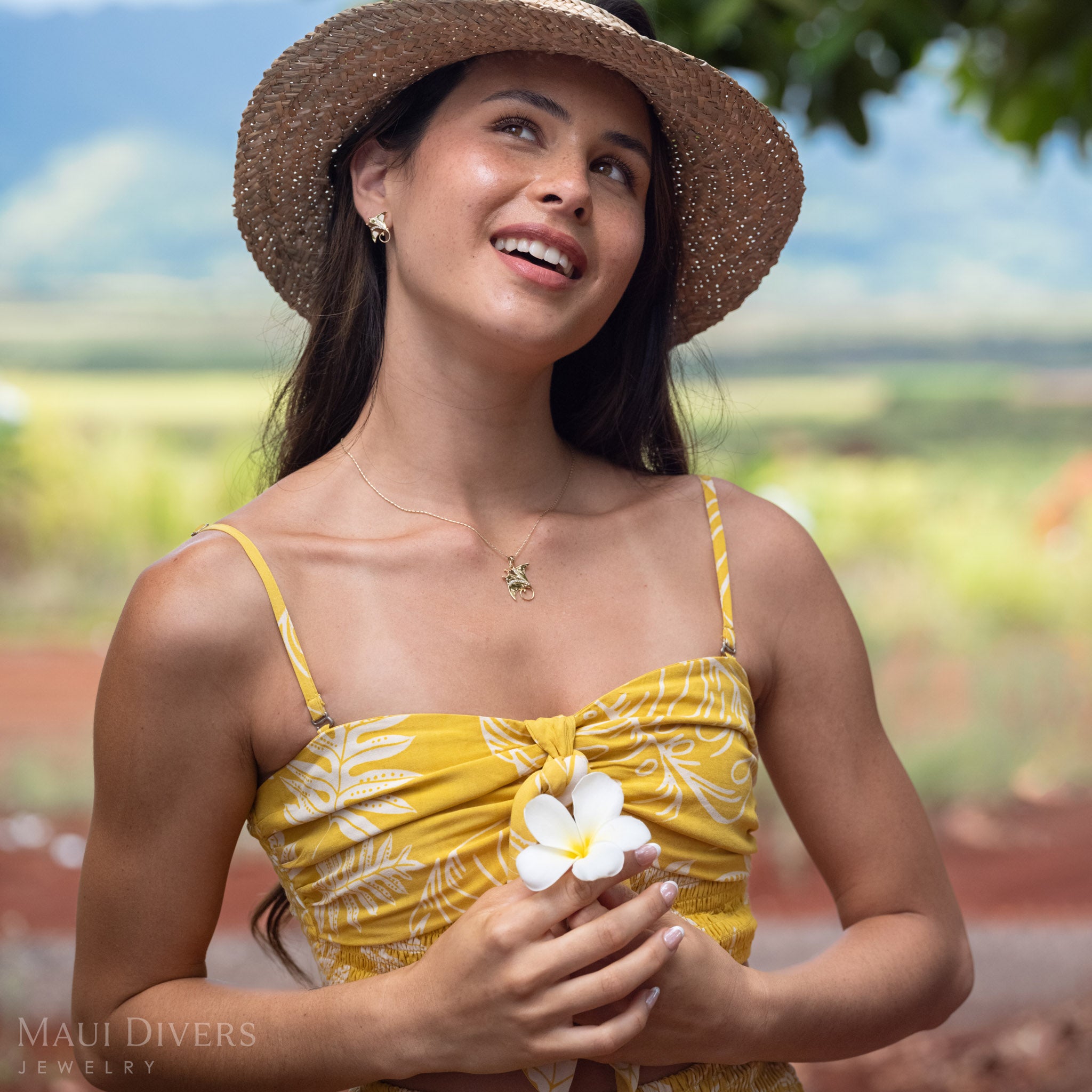 Woman looking up wearing a 14k Gold Manta Ray Pendant around her neck with Natural Diamond detailing and matching earrings holding a white plumeria flower and wearing a brown woven hat. 
