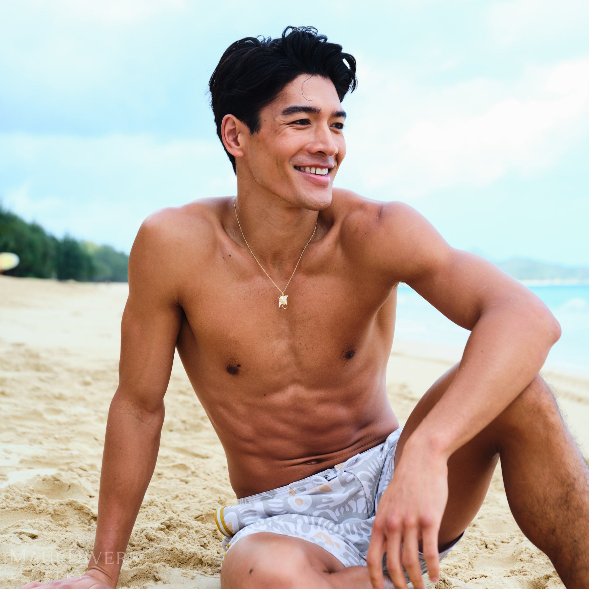 Man sitting on a beach with a clear sky and ocean in the background wearing a gold manta ray pendant