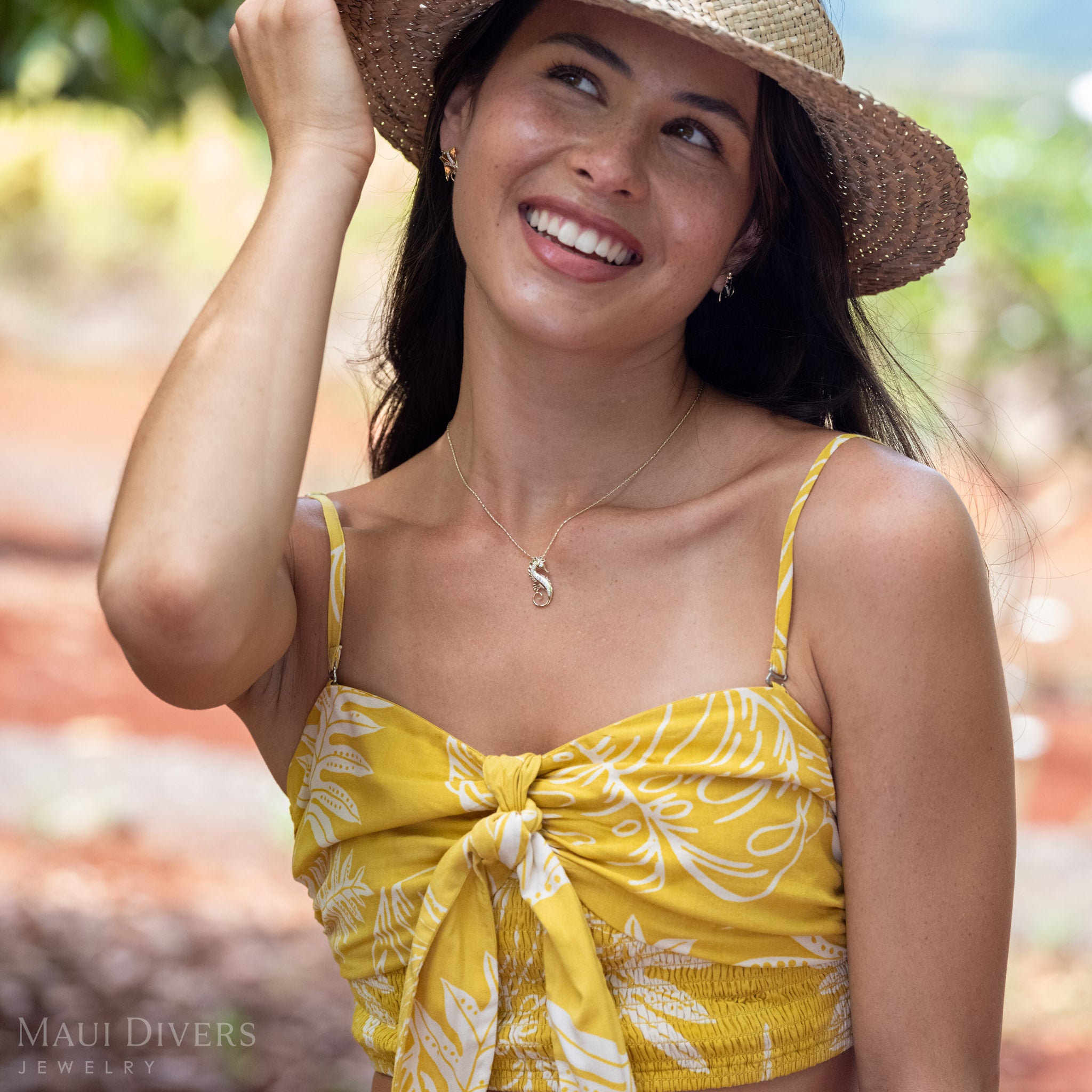 Woman smiling wearing a 14k Gold Seahorse Necklace with Natural Diamond detailing and Manta Ray Gold Earrings with a brown woven hat. 
