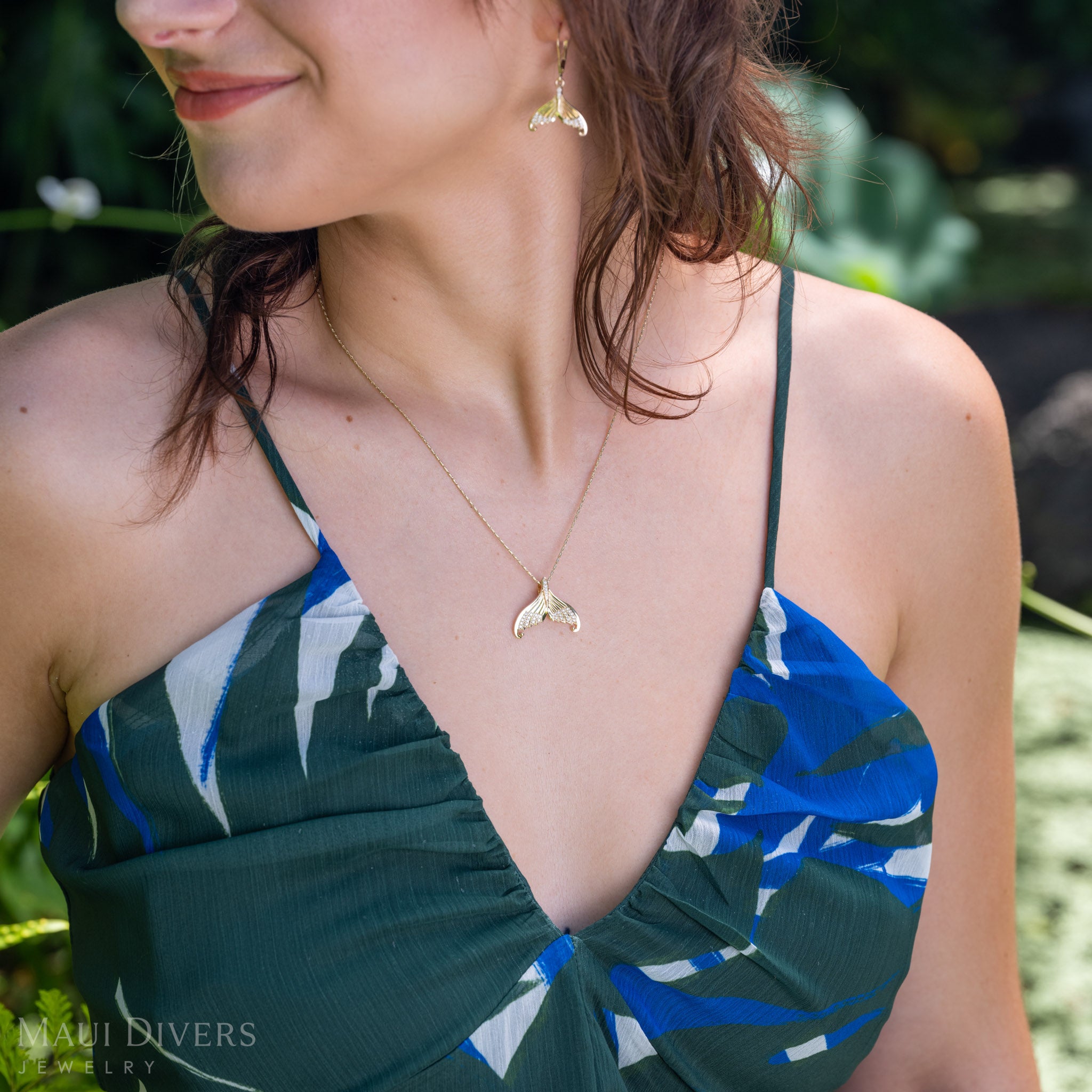 A close-up lifestyle shot of a woman wearing a gold and diamond mermaid tail necklace and matching gold earring, paired with a blue, green, and white patterned halter top. The background is a soft-focus green outdoor setting.