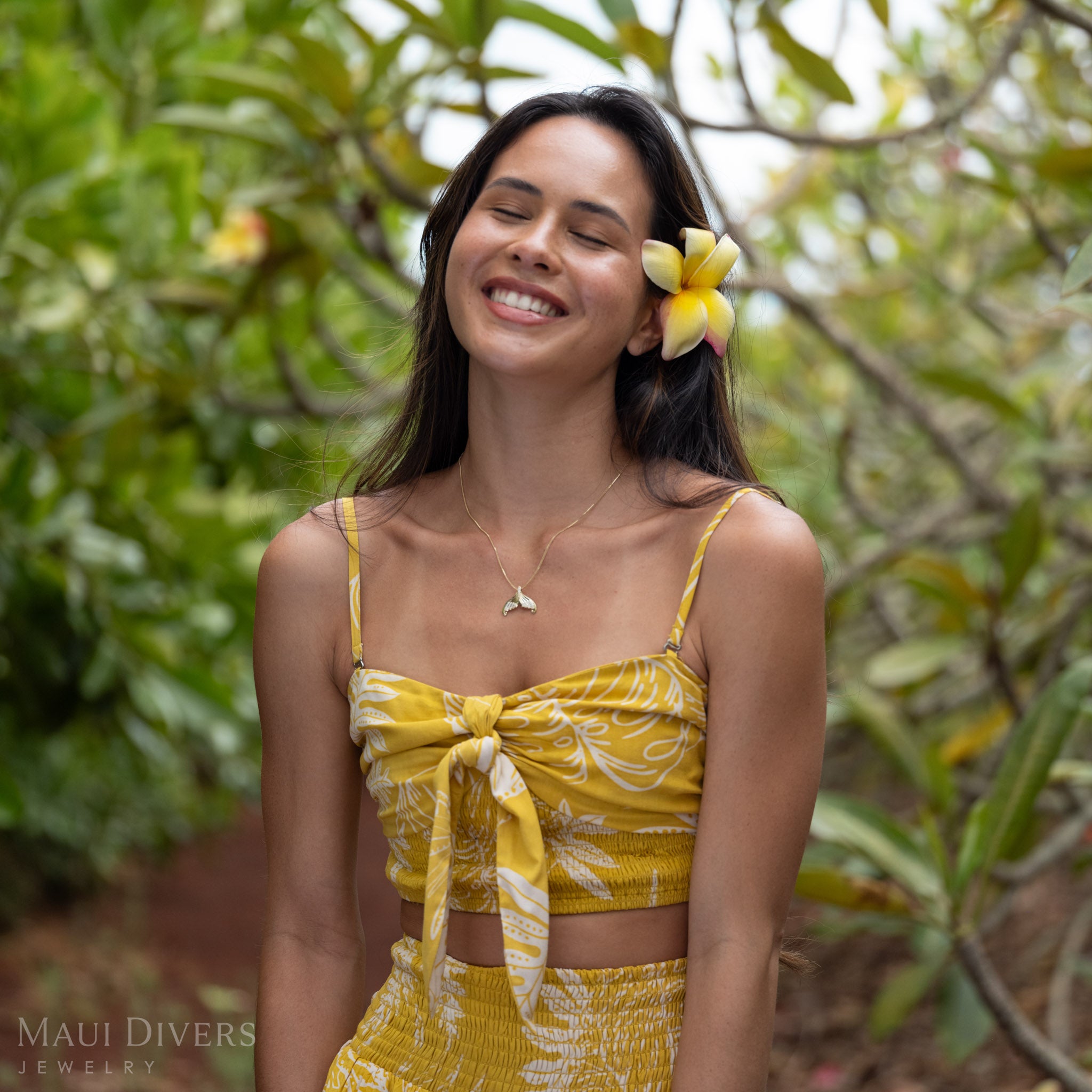 A woman smiling wearing a Yellow Gold Mermaid Tail Pendant with Natural Diamond accents and a yellow plumeria flower in her hair against blurry plumeria trees in the background.