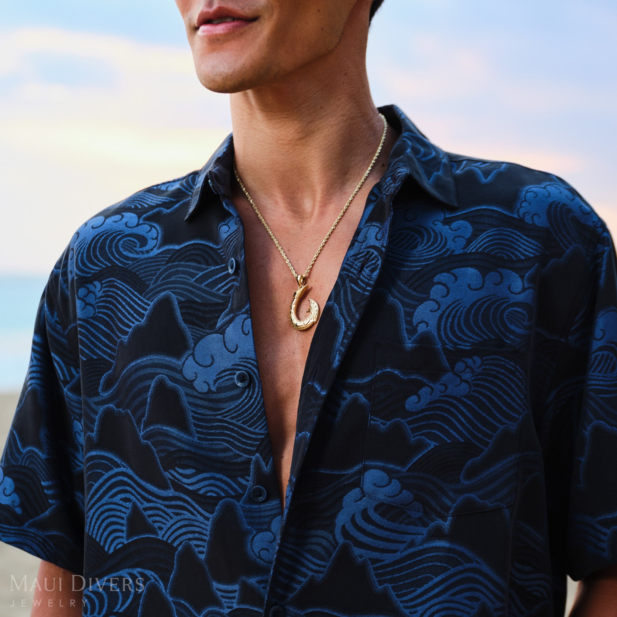 Man wearing a Hawaiian Fish Hook Pendant and blue patterned shirt with a necklace, outdoors.