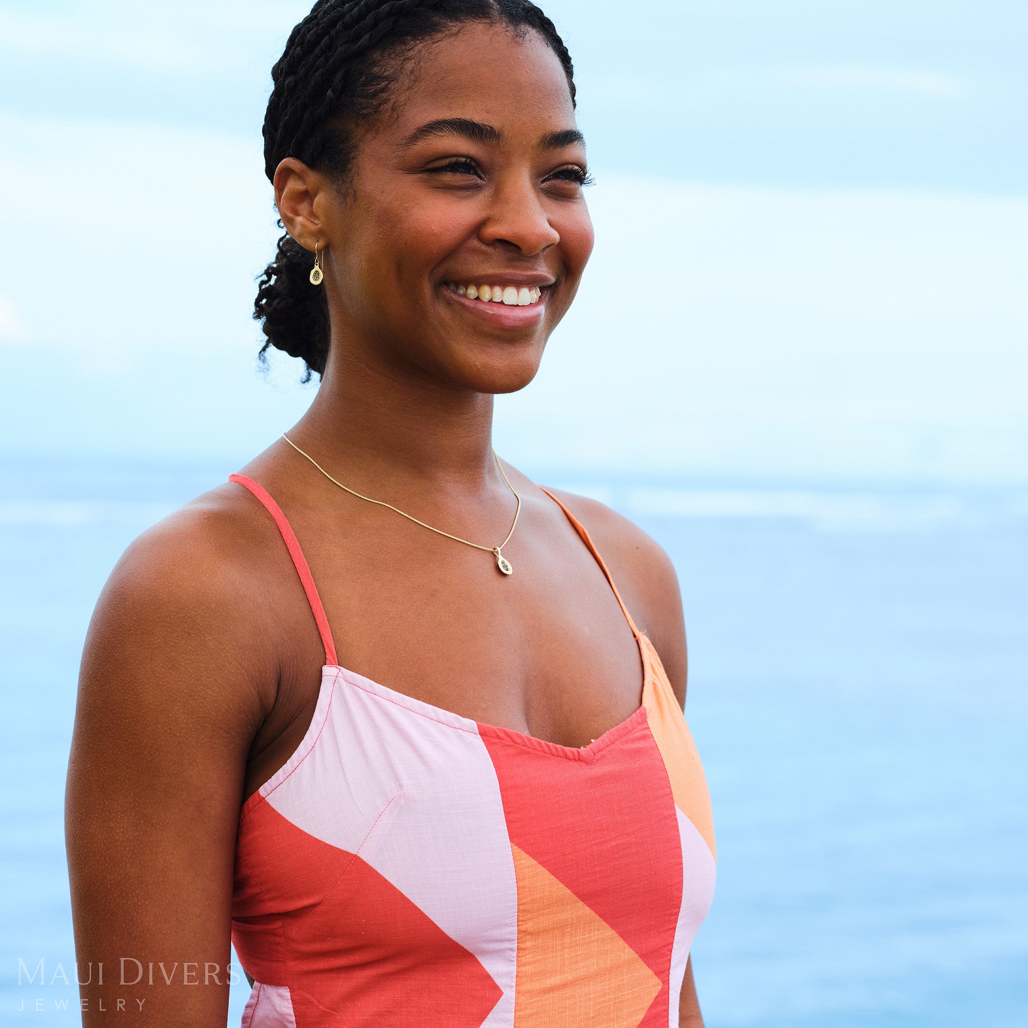 Smiling woman in a pink and orange dress wearing a Cute Fruits Papaya Earring and matching Pendant, both with black diamonds in 14k yellow gold, against a blurred ocean background
