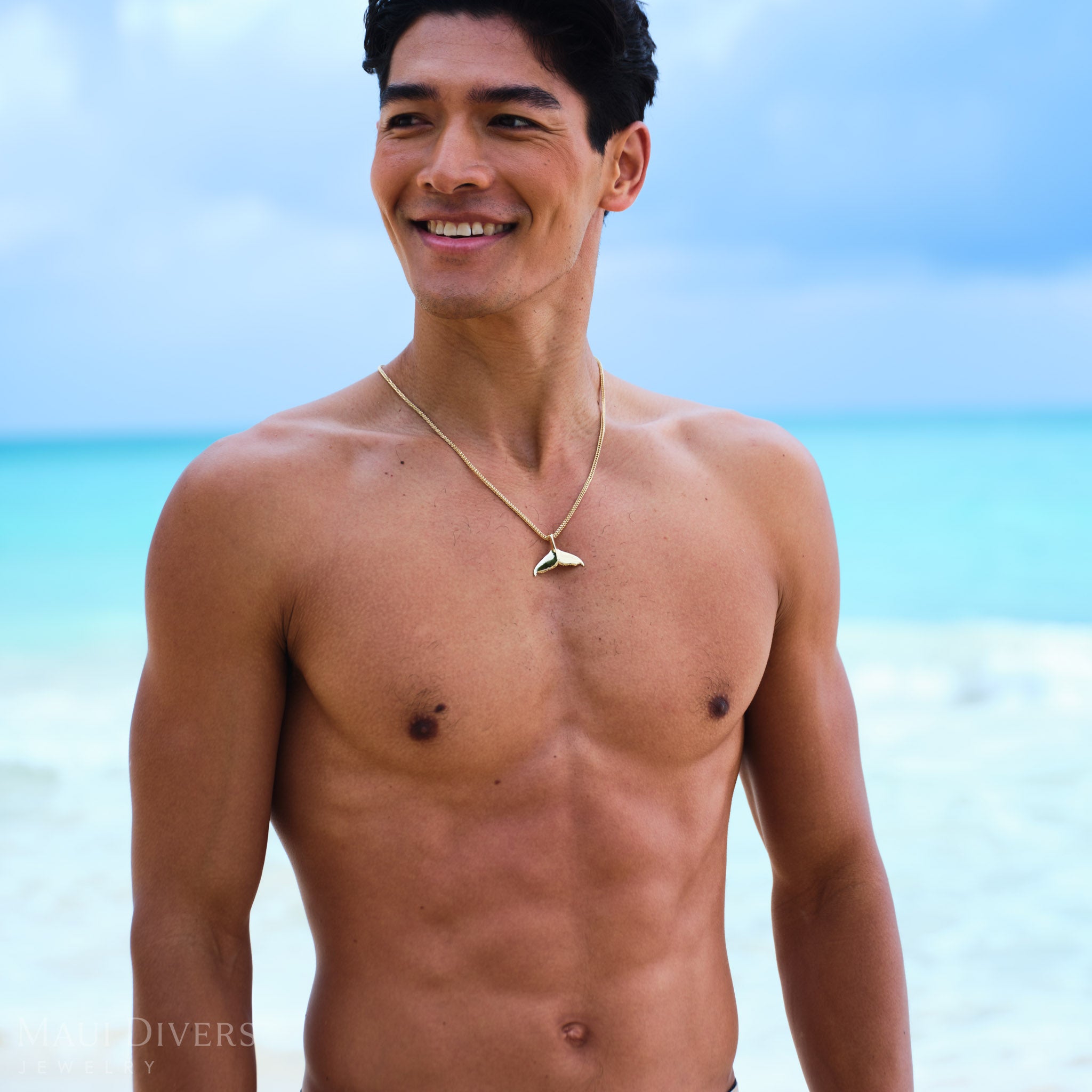 Man standing on a beach wearing a whale tail pendant in gold with clear blue water and sky