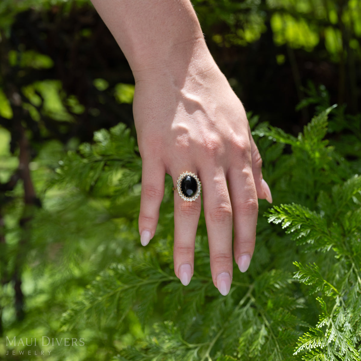 A hand wearing a yellow gold ring with a large oval Hawaiian black coral cabochon and a halo of diamonds, set against a background of green fern foliage. 
