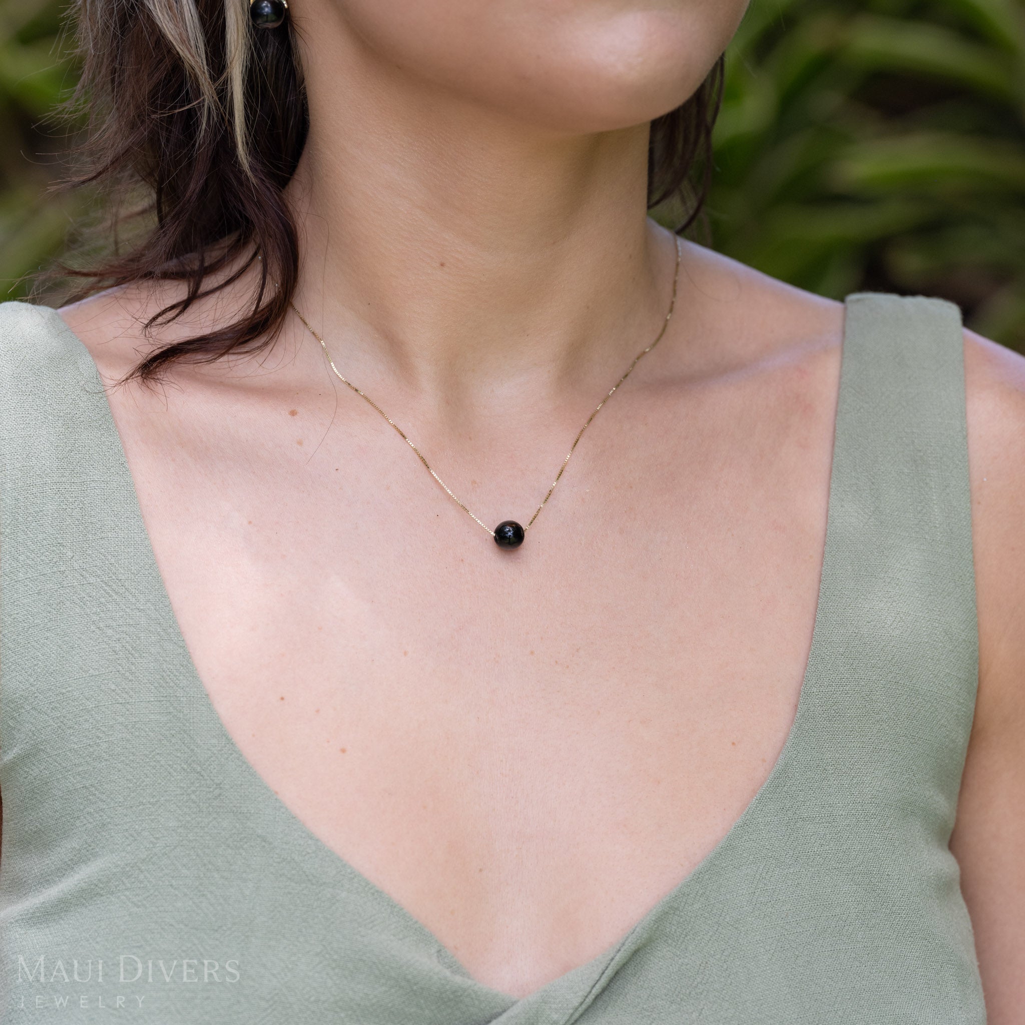 A close-up of a woman in a light green top wearing an Adjustable Black Coral Slider Necklace in 14k yellow gold, against a blurred leafy green background