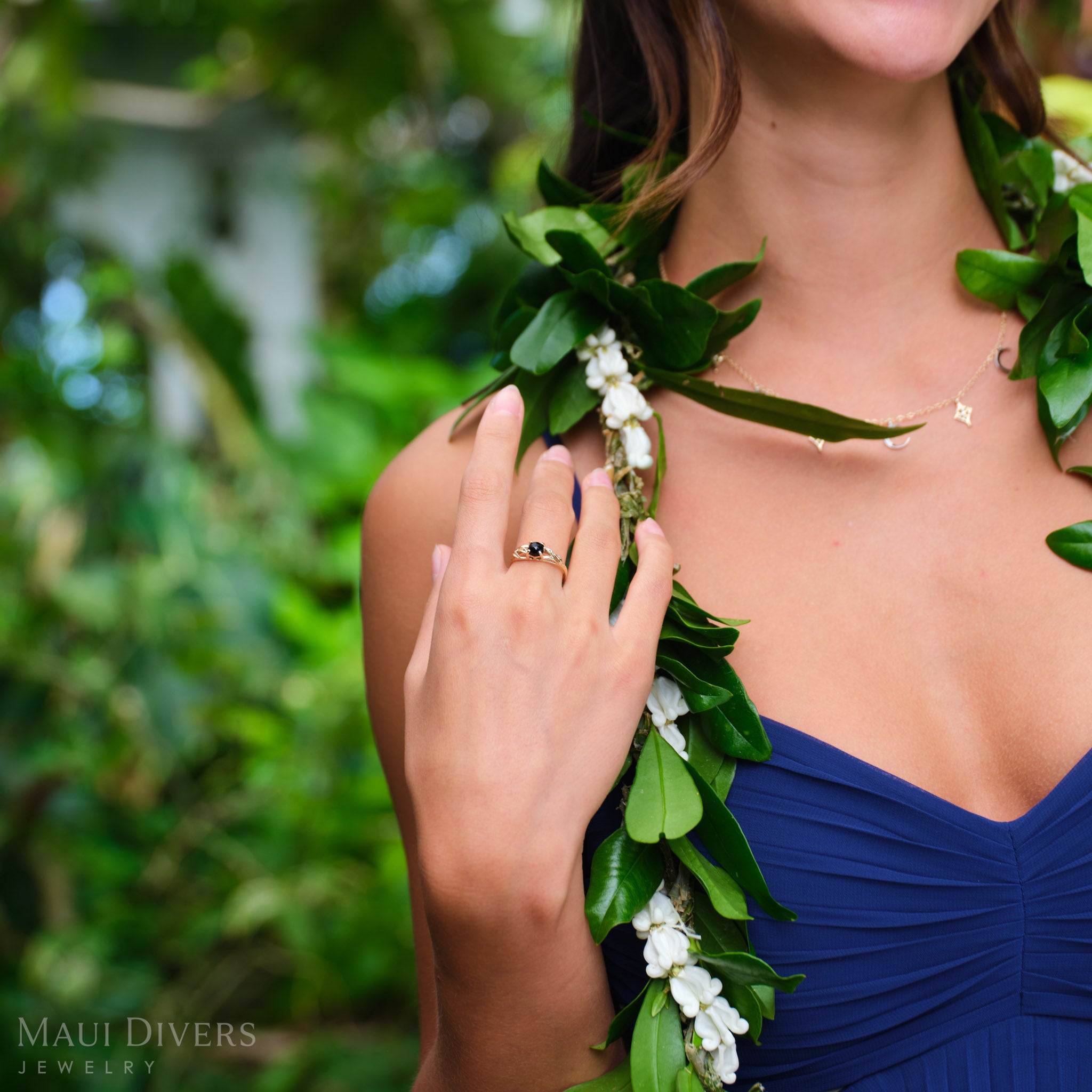 Woman in a blue top and maile lei wearing a Maile Leaf Black Coral Ring in 14k yellow gold on her hand, against a blurred leafy green background