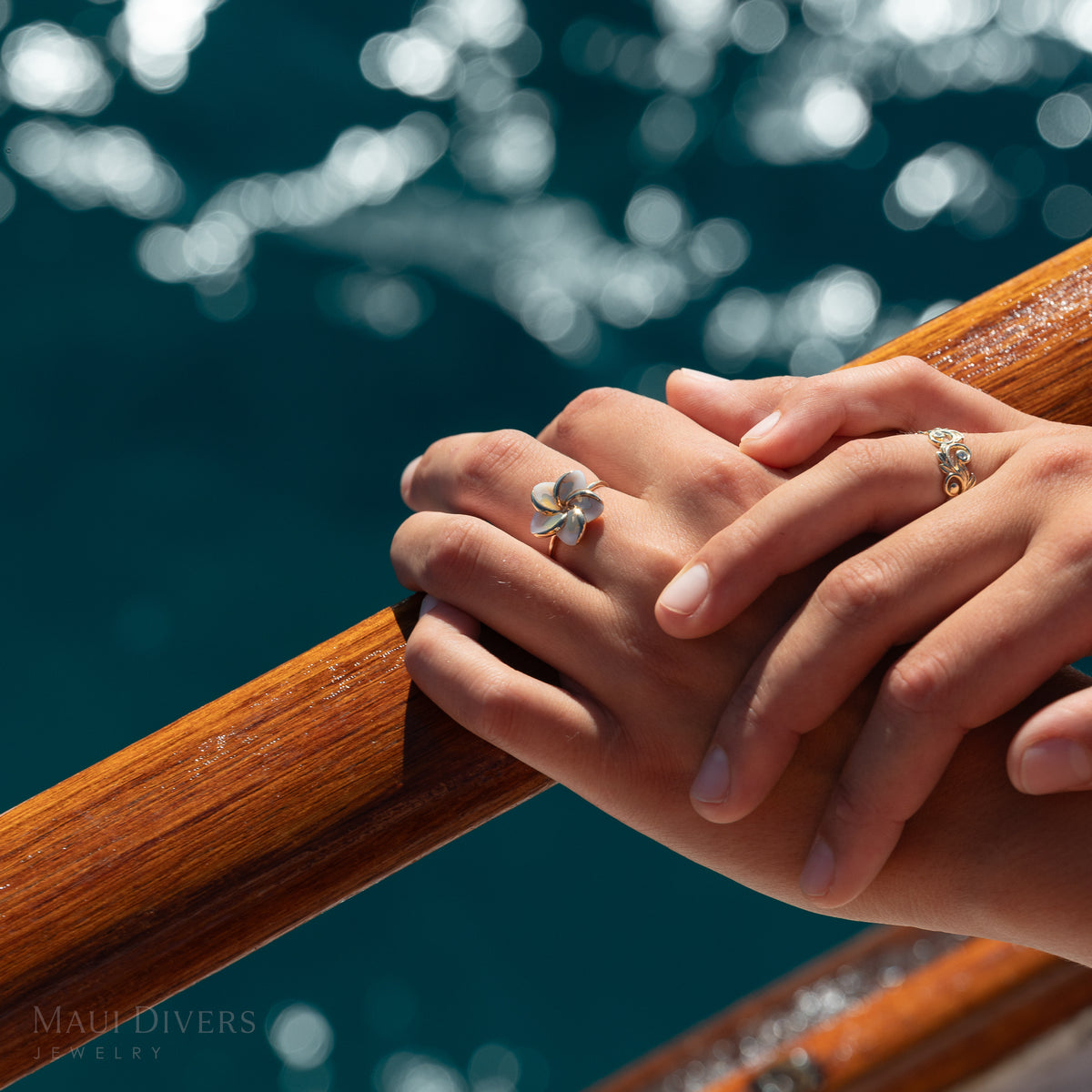 Close-up of hands resting on a wooden rail wearing a gold plumeria mother of pearl ring and a intricate filigree ring, against a sparkling deep blue ocean background