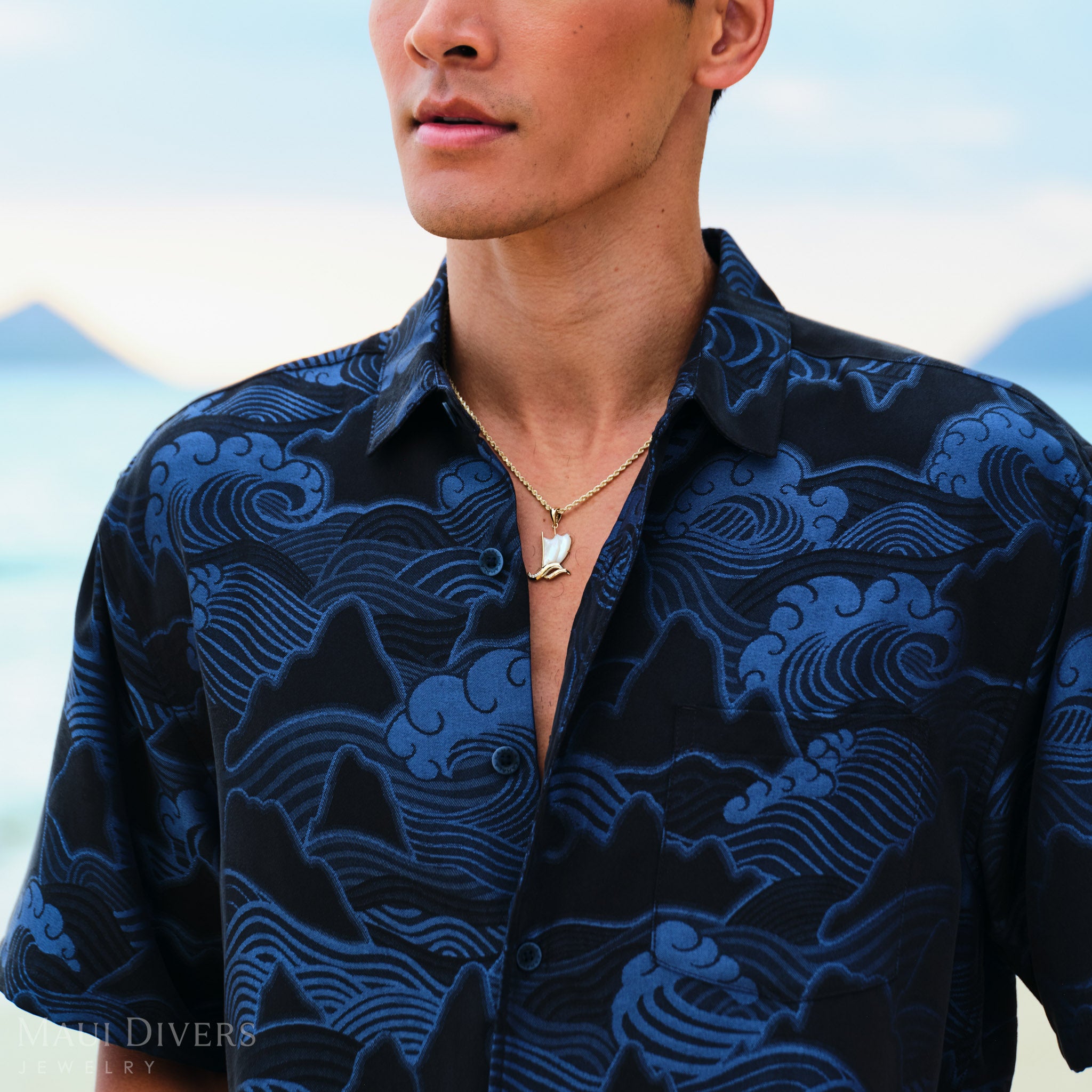 Waʻa (Canoe) Pendant on a gold chain worn by a man in a blue patterned shirt by the ocean