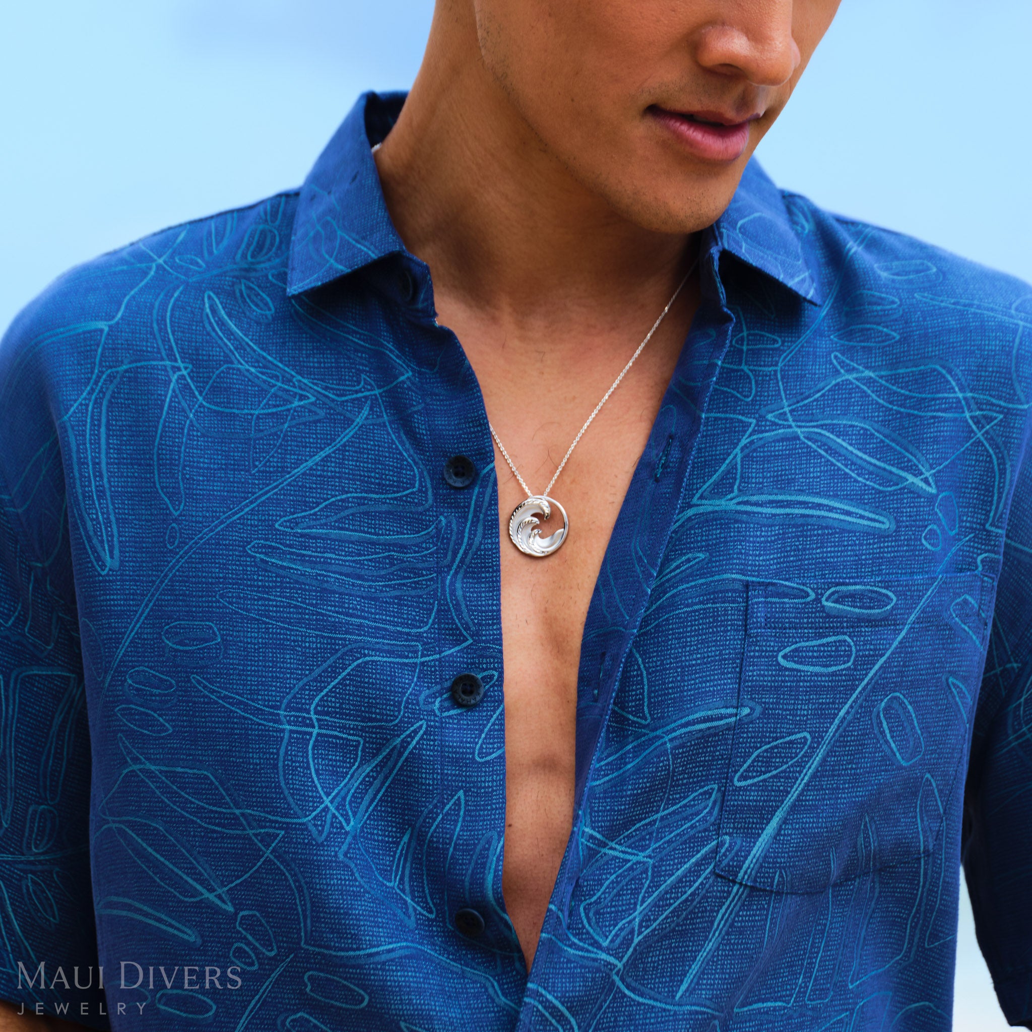 Man wearing a blue patterned shirt with a round silver wave necklace 