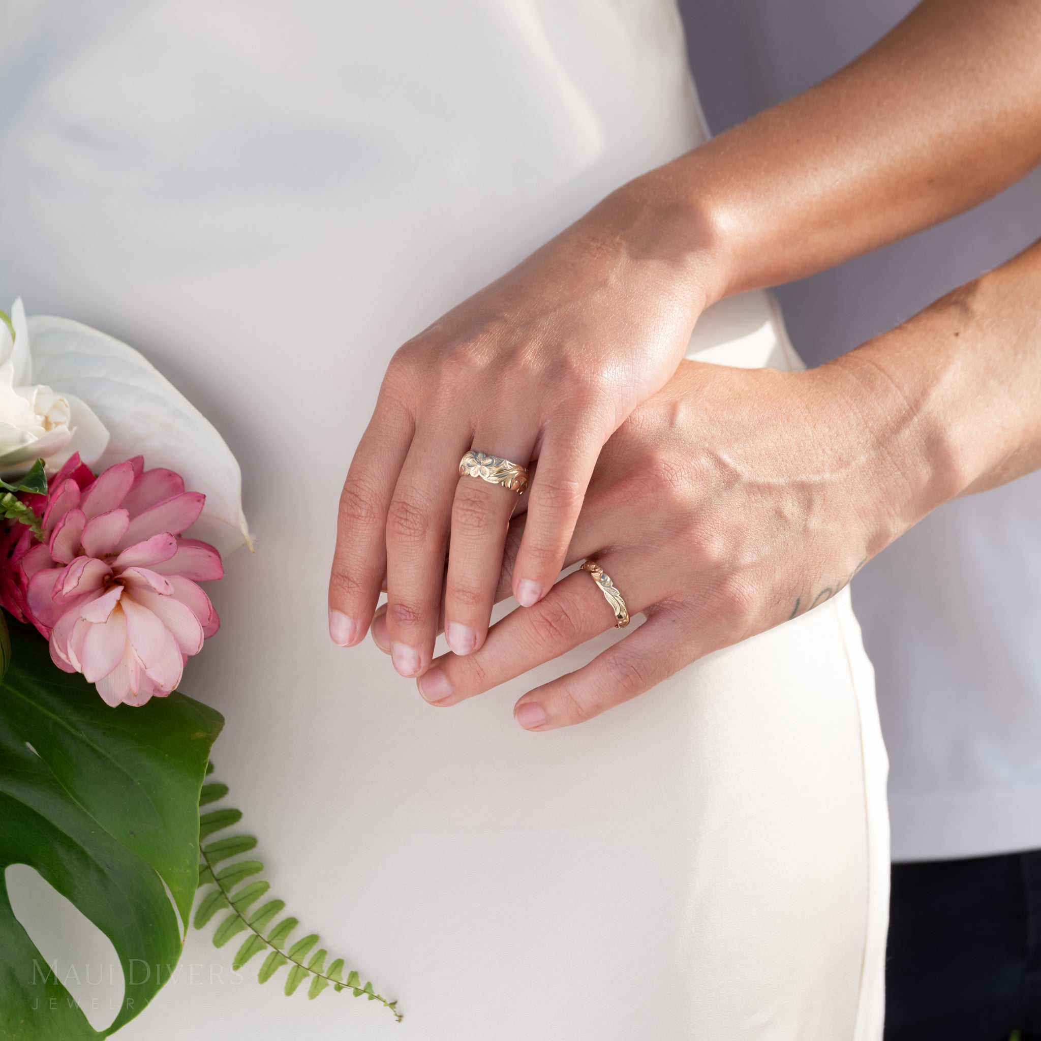 Close-up of a woman and man holding hands wearing an 8mm Hawaiian Heirloom Plumeria Scroll Ring and a Hawaiian Heirloom Old English Scroll Ring over a white dress and bouquet on the side