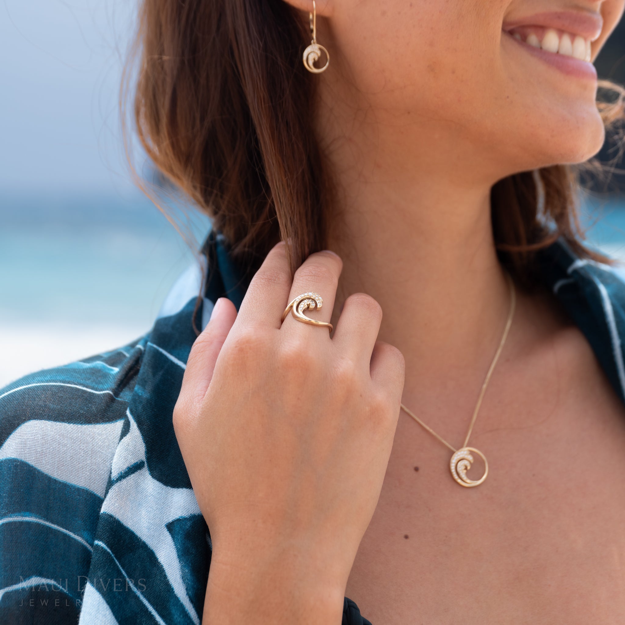 Close-up of a hand wearing a gold and diamond Nalu (wave) ring with a blurry beach in the background