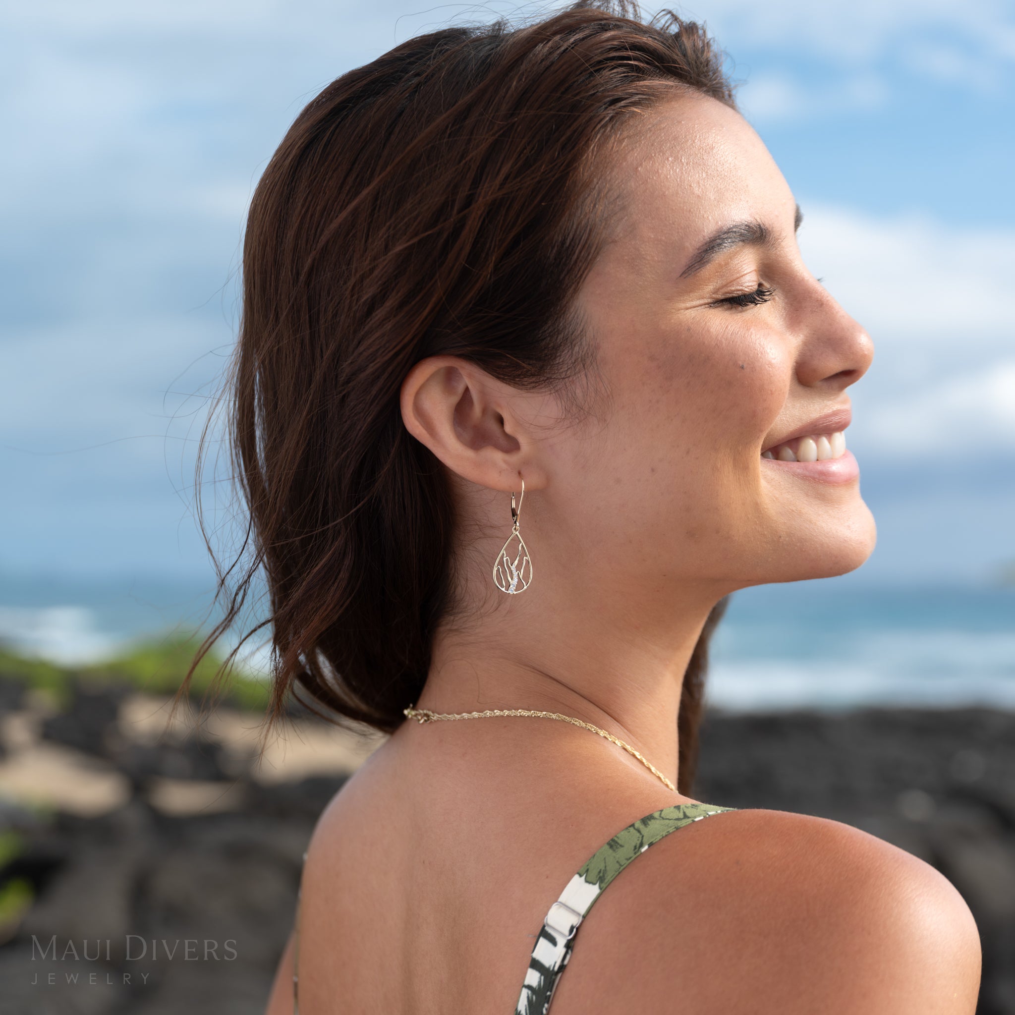 Smiling woman wearing a gold and diamond Koʻolau waterfall teardrop earring with a blurry blue sky and dark rocks in the background