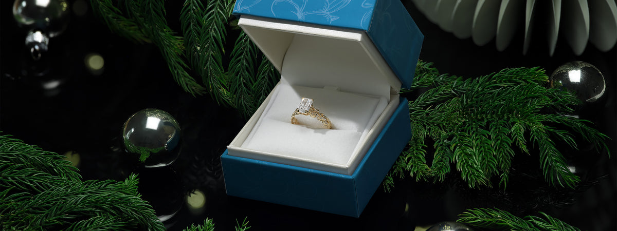 A Living Heirloom Collection engagement ring in gold with a radiant diamond solitaire, displayed inside an open blue jewelry box on a dark festive background decorated with pine branches and glass ornaments.