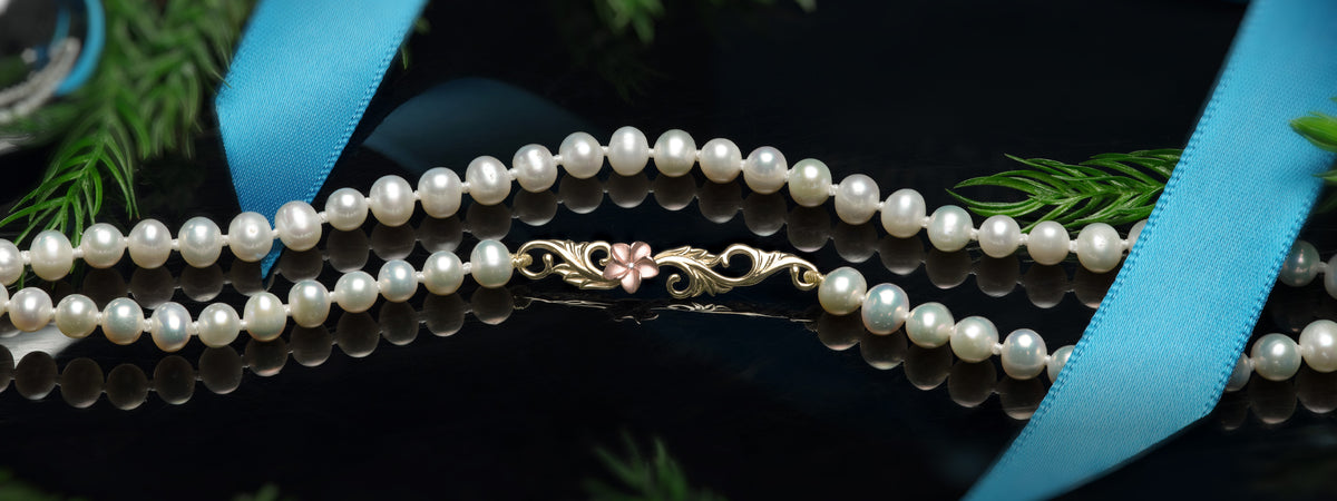 A white freshwater pearl strand with a gold-toned Hawaiian Heirloom clasp with a rose gold plumeria, styled with a light blue ribbon on a reflective black surface.