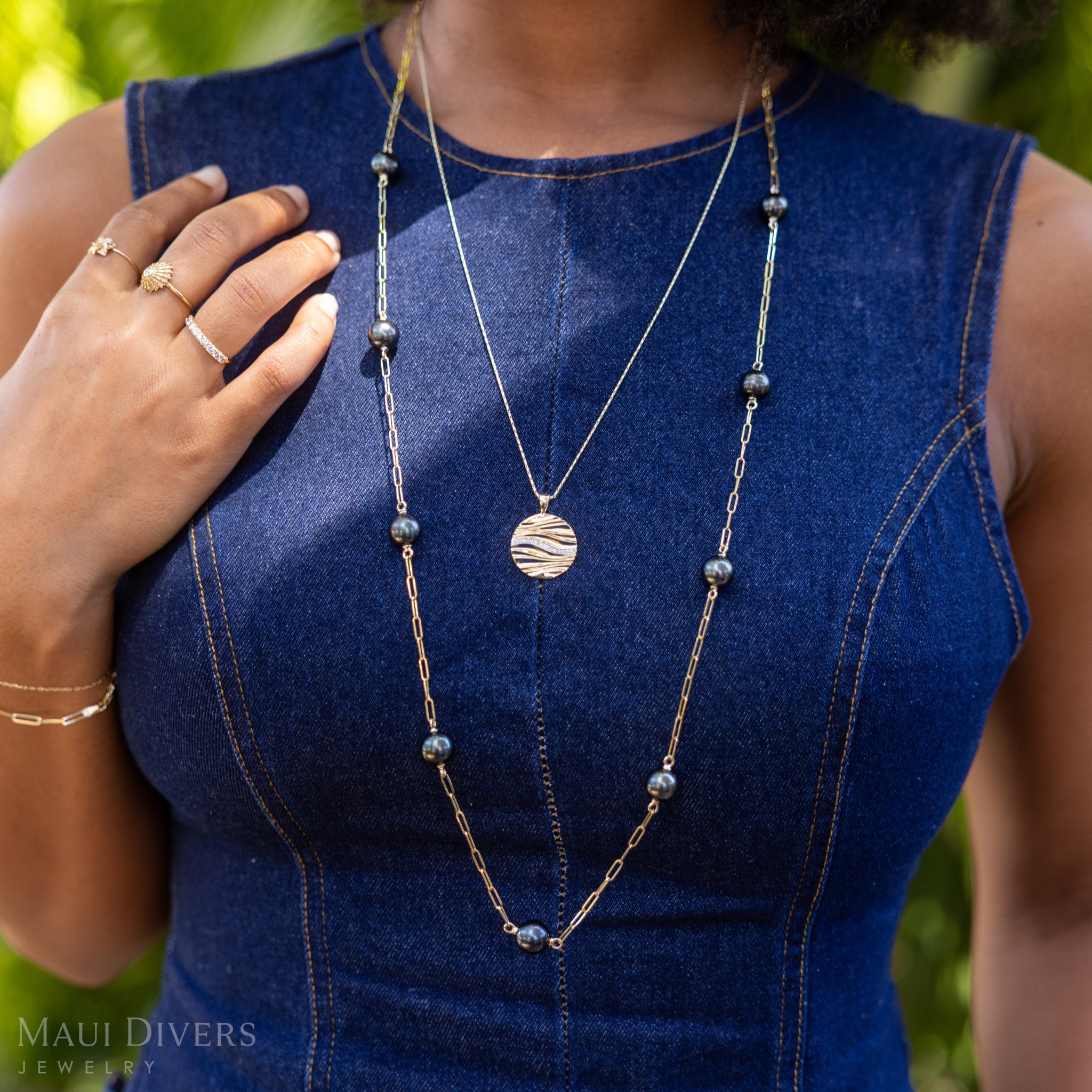 A person wearing a long Tahitian black pearl necklace and the Ocean Sand Pendant with a blue sleeveless top outdoors